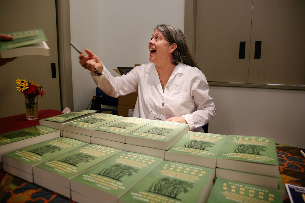 woman in white shirt looking happy to recognize someone as she sits behind a pile of her books
